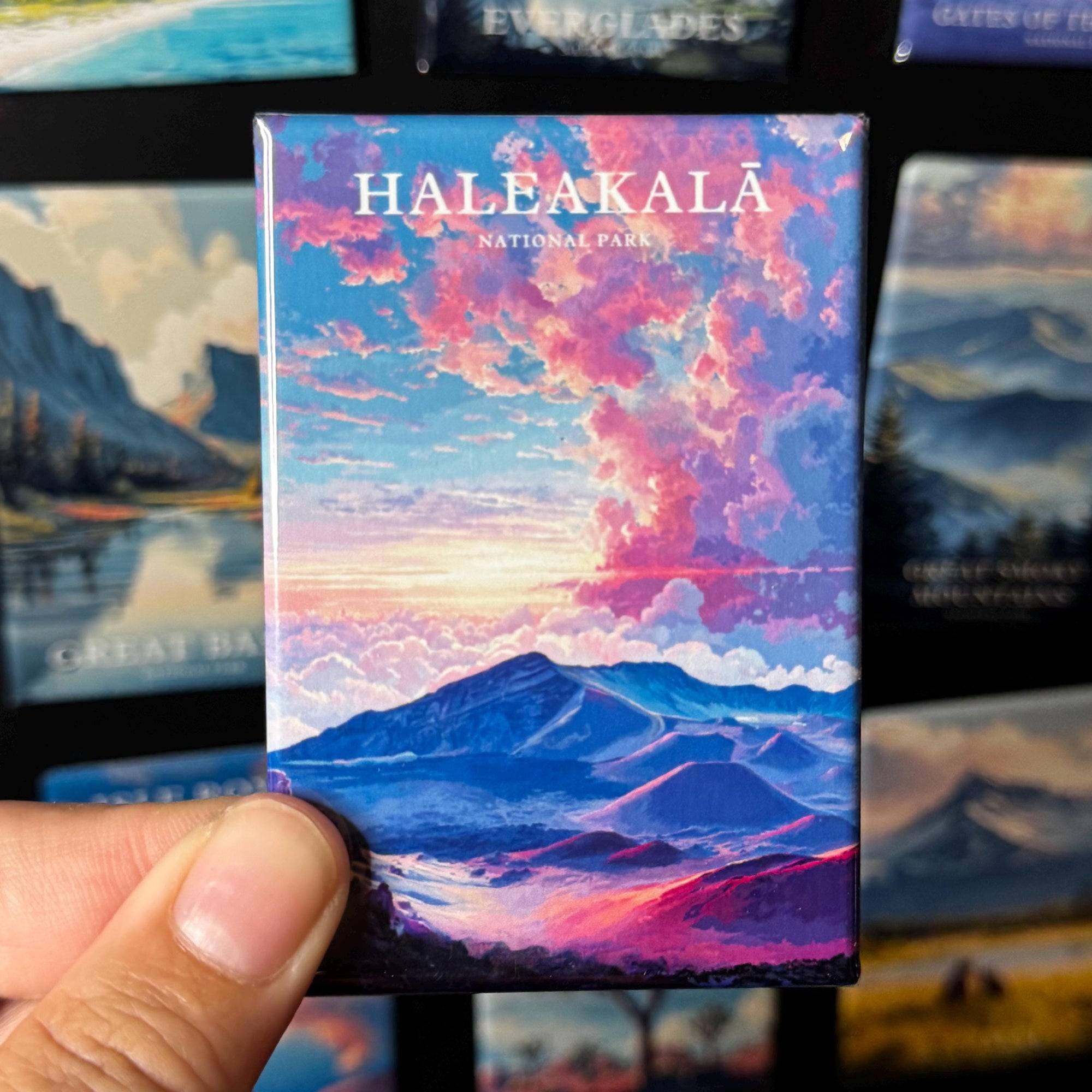 A hand holding a magnet with a colorful landscape depicting a mountain range and a sunset sky.