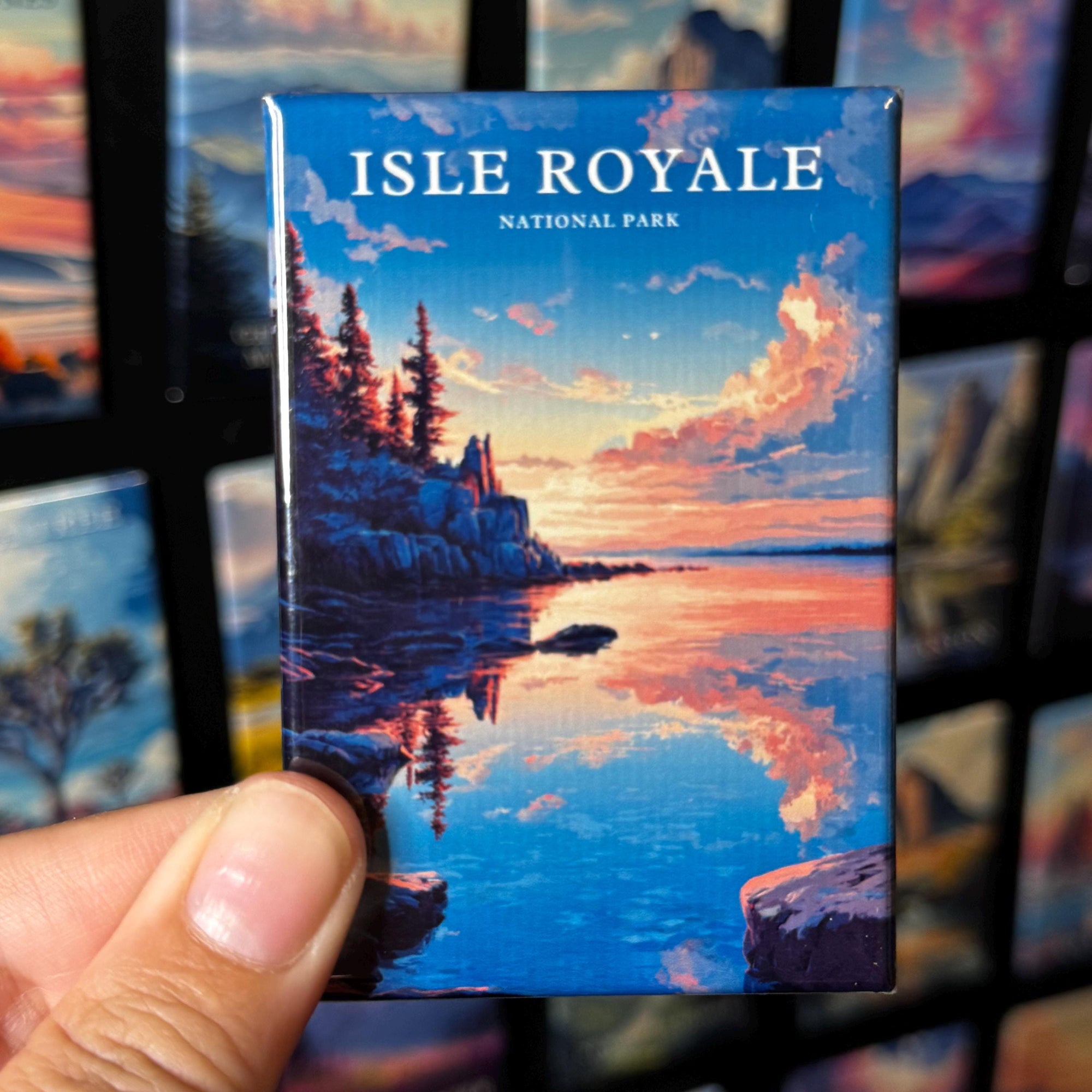 A hand holding a magnet with a scenic image of Isle Royale National Park, featuring a sunset over the water and rocky cliffs.