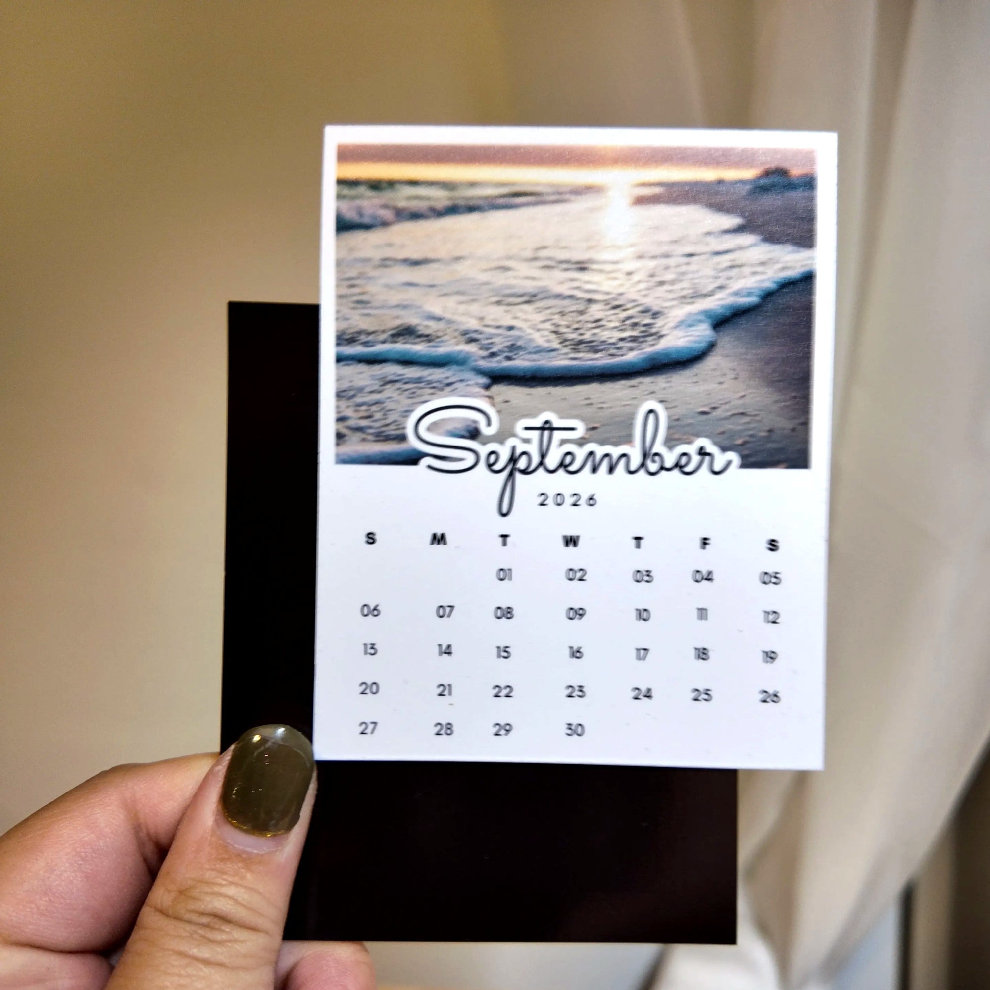 A person's hand holding a calendar card with a beach scene on the cover.
