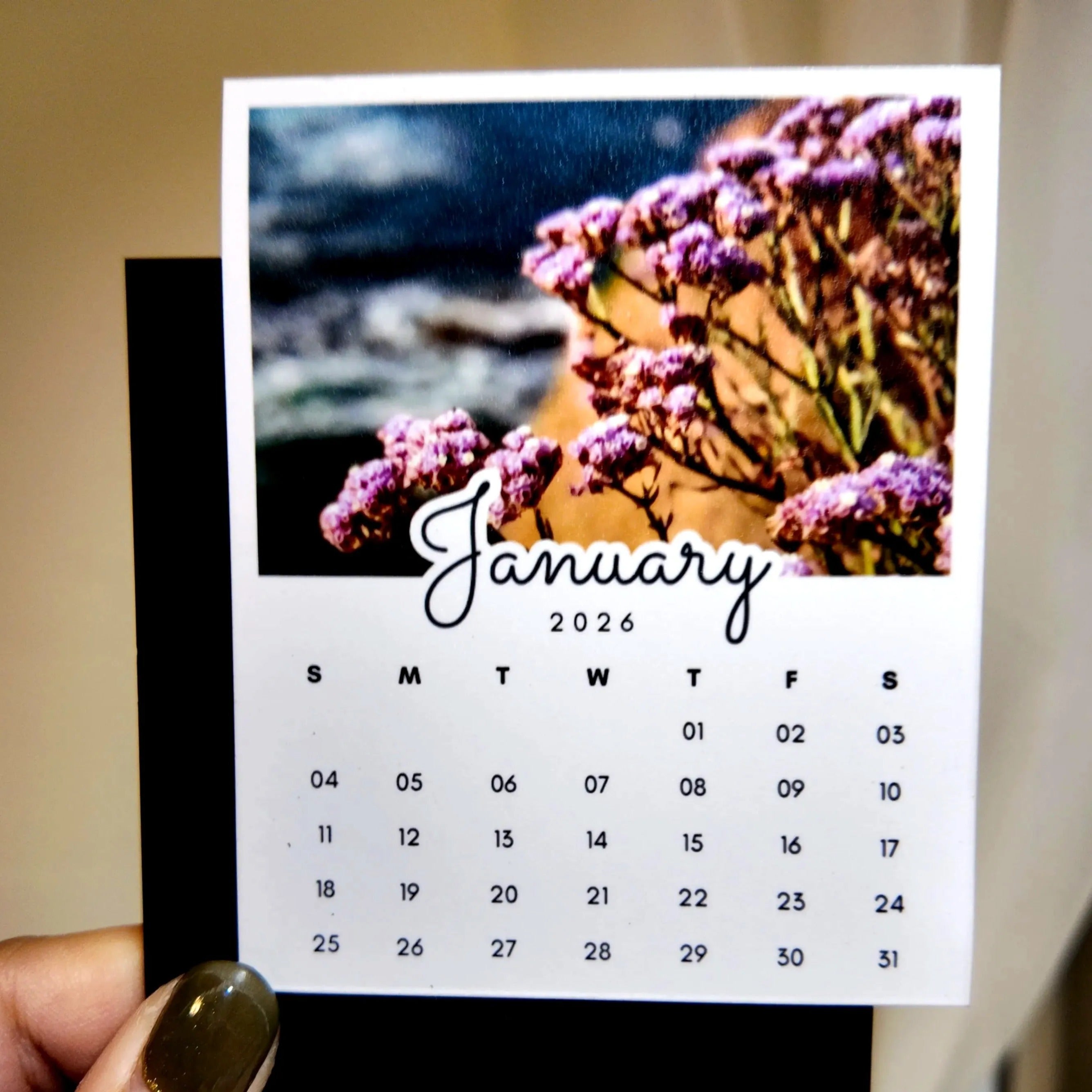 A person's hand holding a calendar card with a photo of purple flowers on the cover.