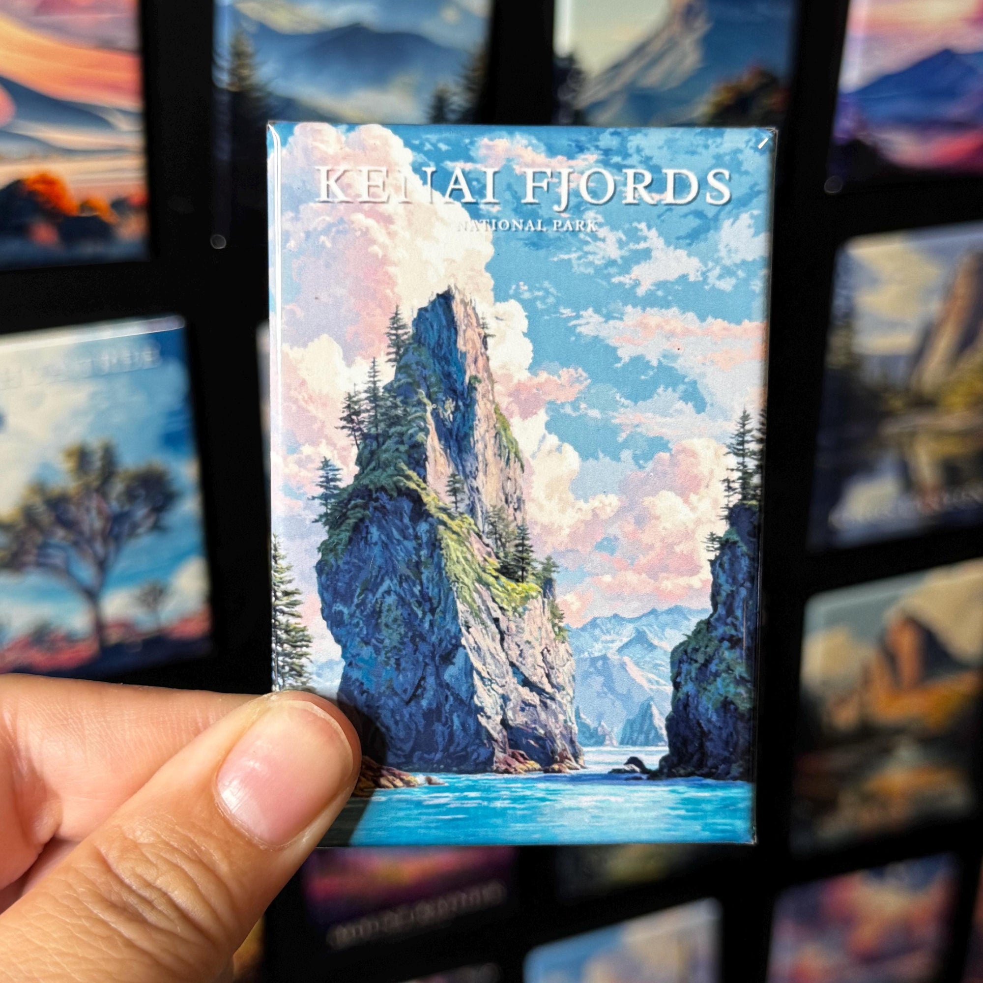 A hand holding a magnet with a scenic mountain landscape, including a river and clouds, against a background of other magnets.