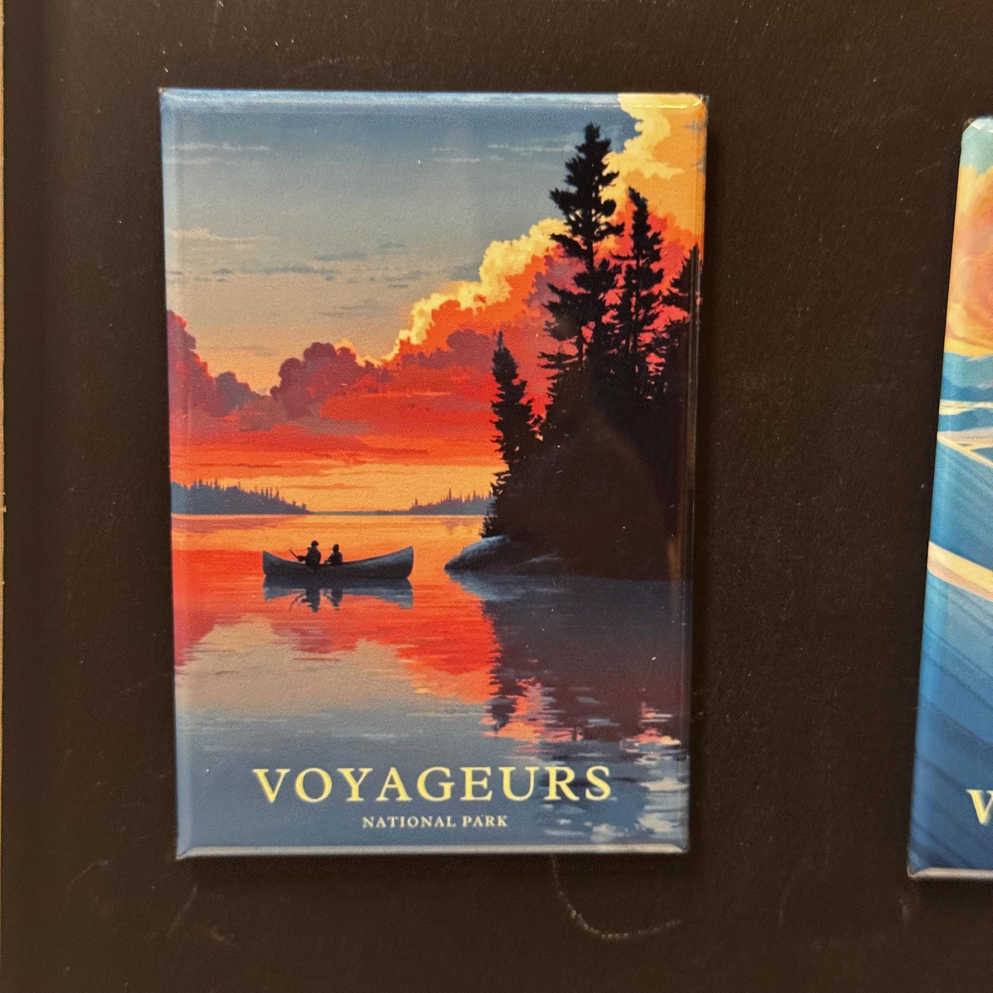 A framed magnet depicting a canoe on a lake with a sunset in the background, surrounded by trees.
