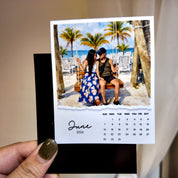 A person is holding a small photo frame containing a printed photo of a couple sitting on a swing, with a beach scene in the background.