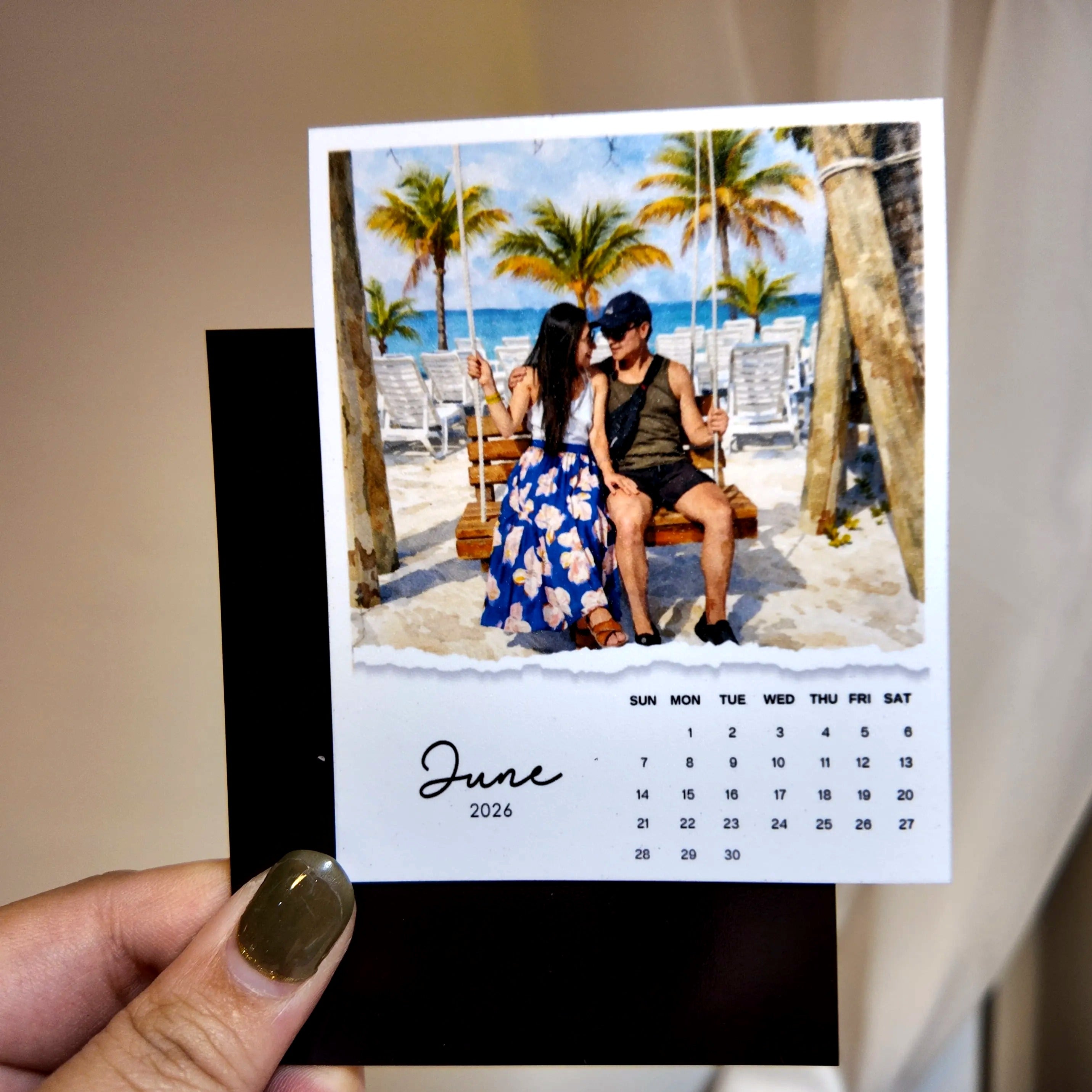 A person is holding a small photo frame containing a printed photo of a couple sitting on a swing, with a beach scene in the background.
