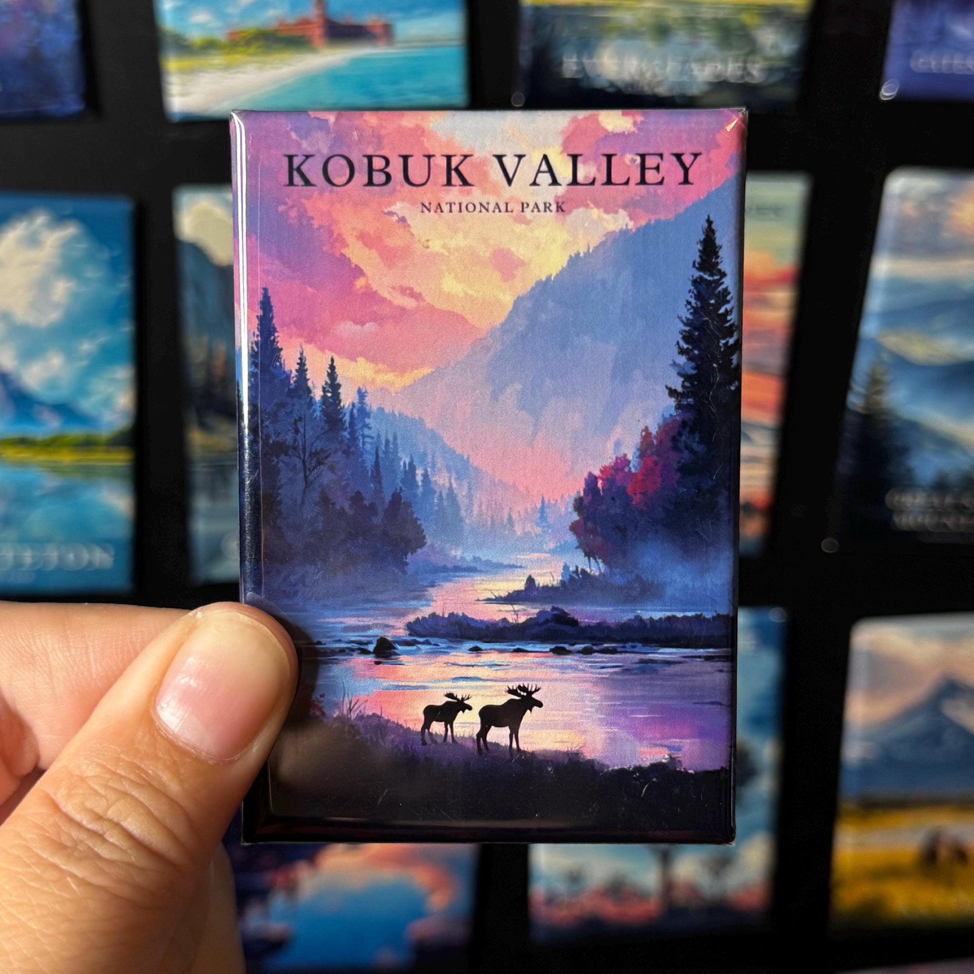 A hand holding a magnet with a scenic landscape of Kobuk Valley National Park, featuring silhouettes of moose and trees against a colorful sunset sky.