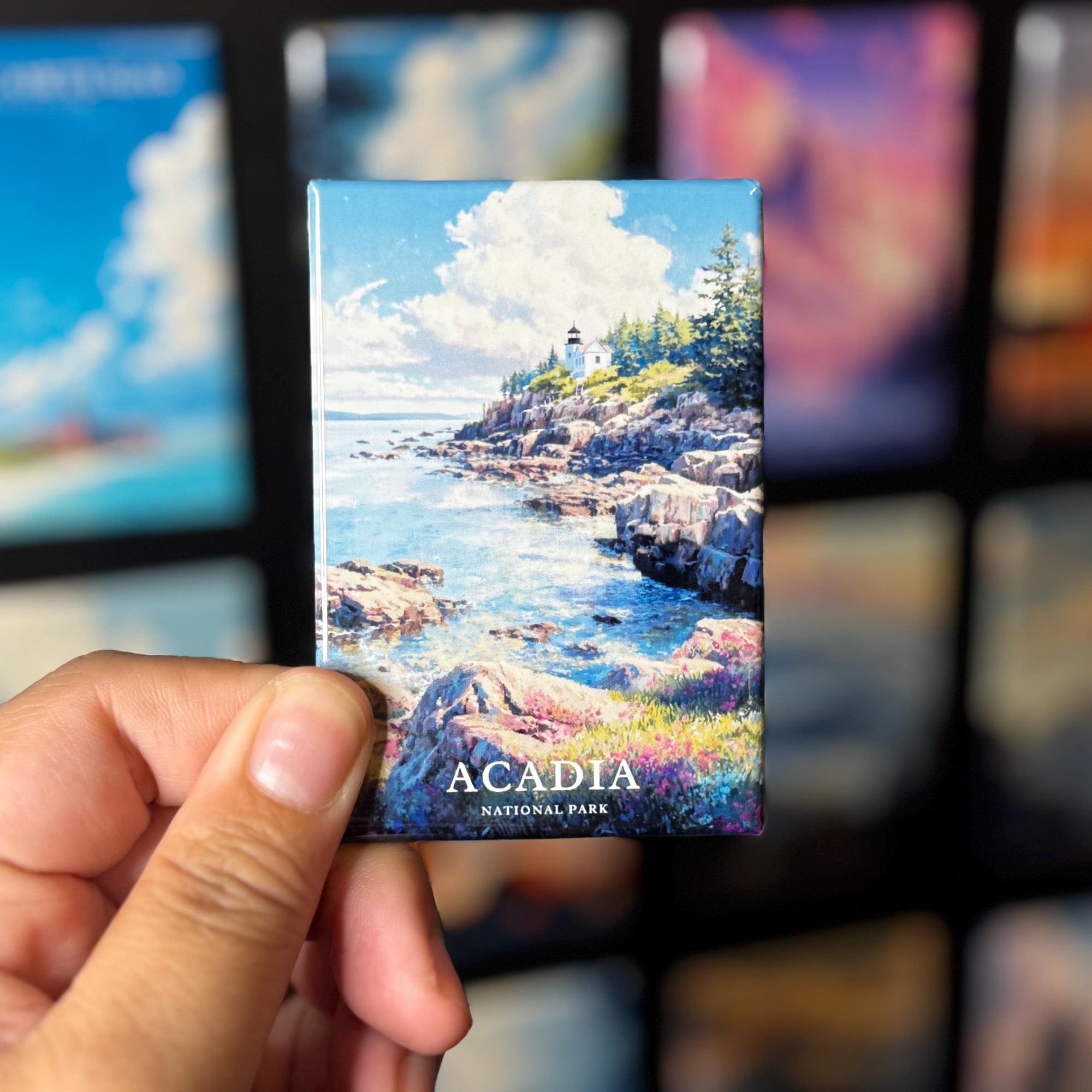A hand holding a magnet with a scenic image of Acadia National Park, featuring a lighthouse and a rocky coastline with water.