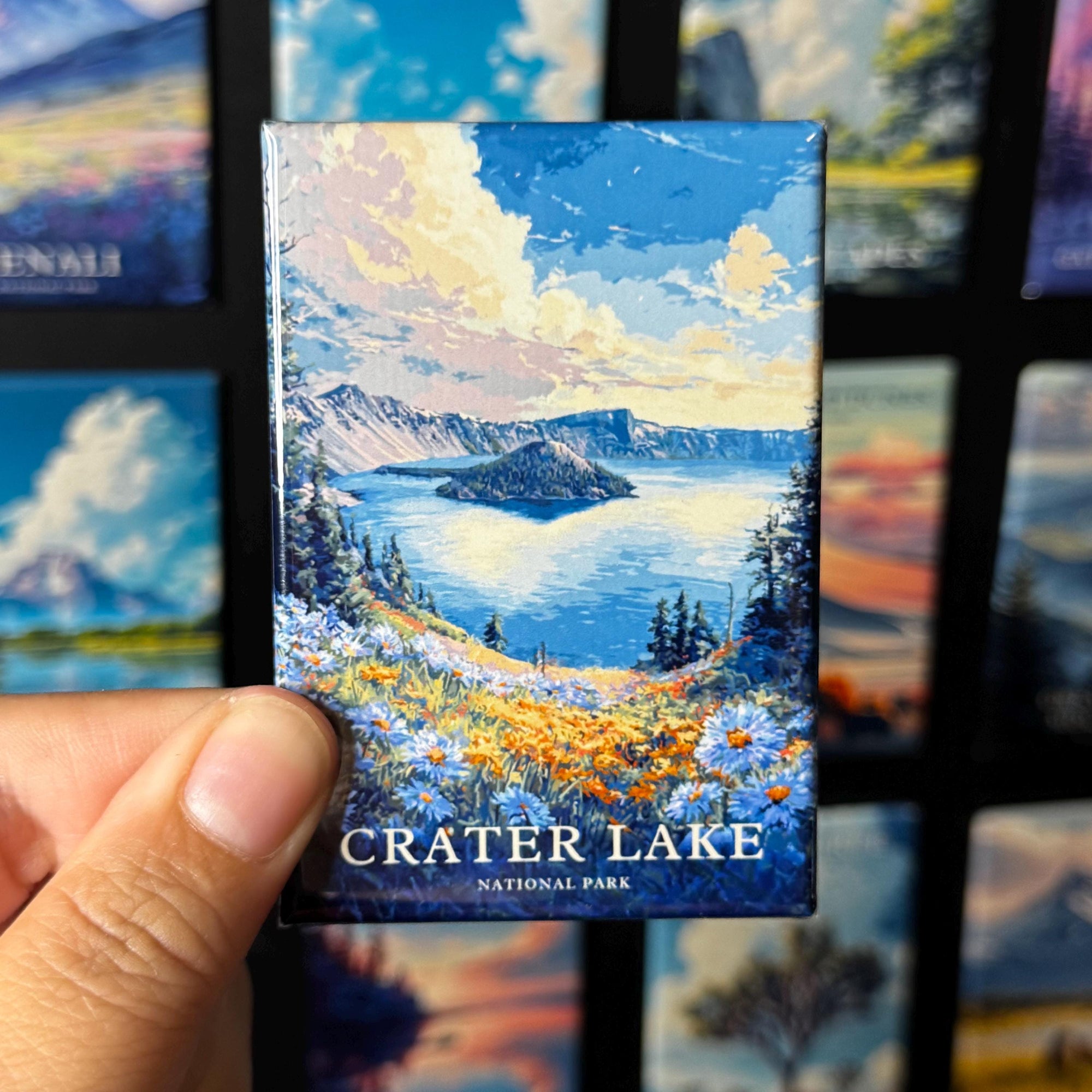 A hand holding a magnet with a painting of Crater Lake in National Park.