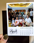 A person is holding a calendar featuring a group photo of a family or group of people, with the date "April 2026" displayed.
