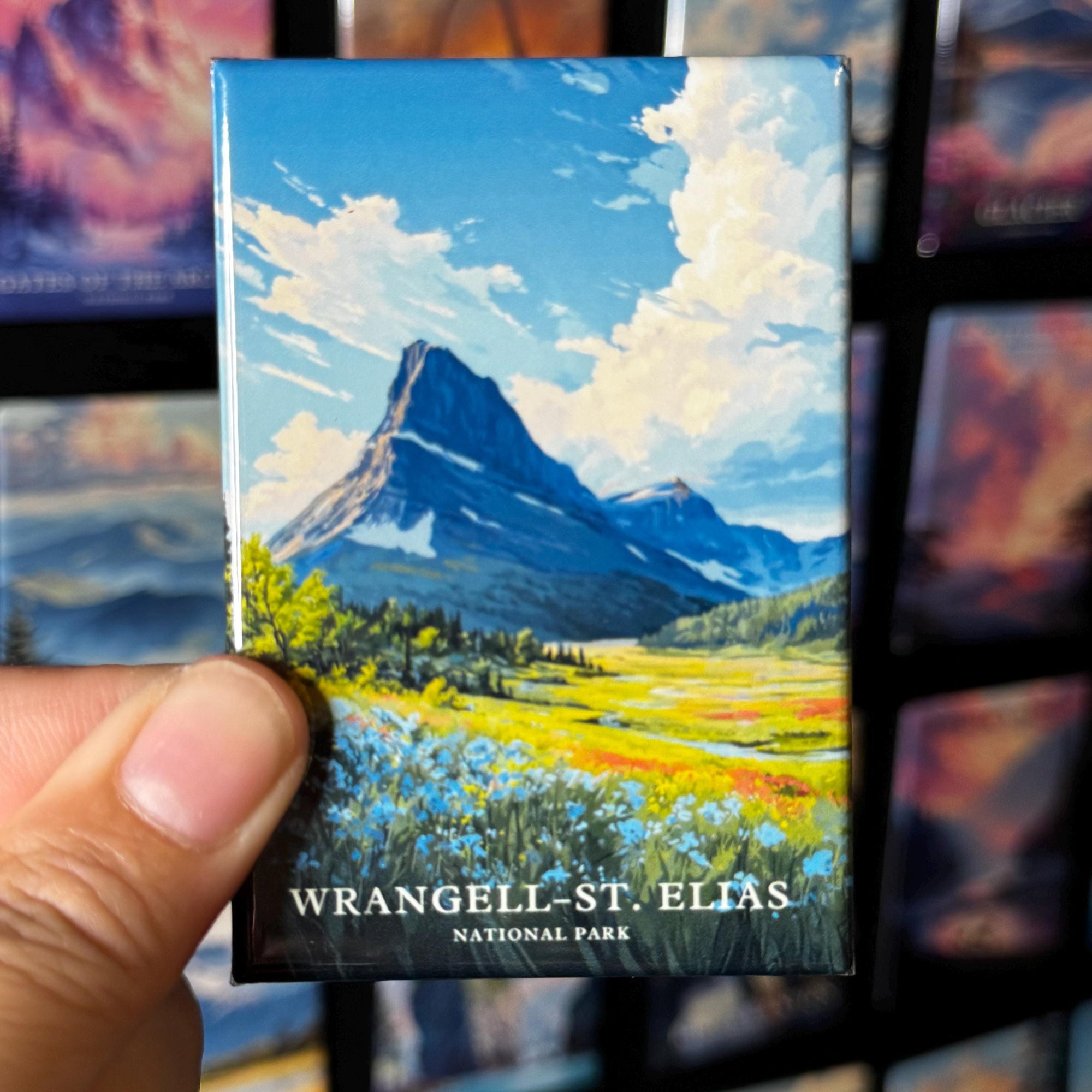 A hand holding a magnet with a painting of a mountain landscape, specifically Wrangell-St. Elias National Park.
