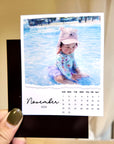 A hand holding a small calendar with a photo of a child sitting in a pool.