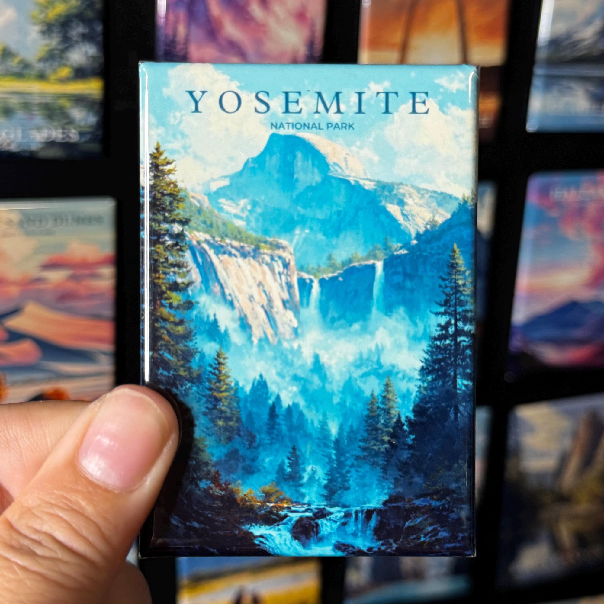 A hand holding a magnet with a scenic image of Yosemite National Park, featuring a waterfall and forest.