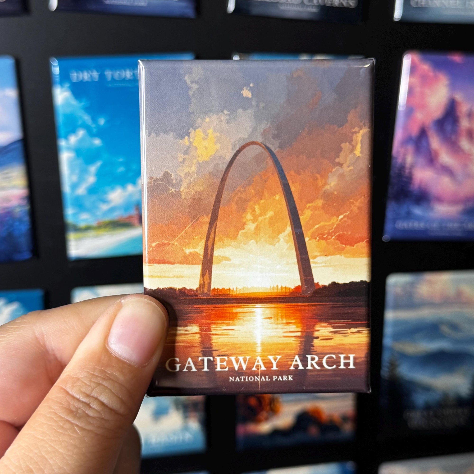 A person's hand holding a magnet with an illustration of the Gateway Arch, a famous monument in Gateway National Park, with a sunset in the background.