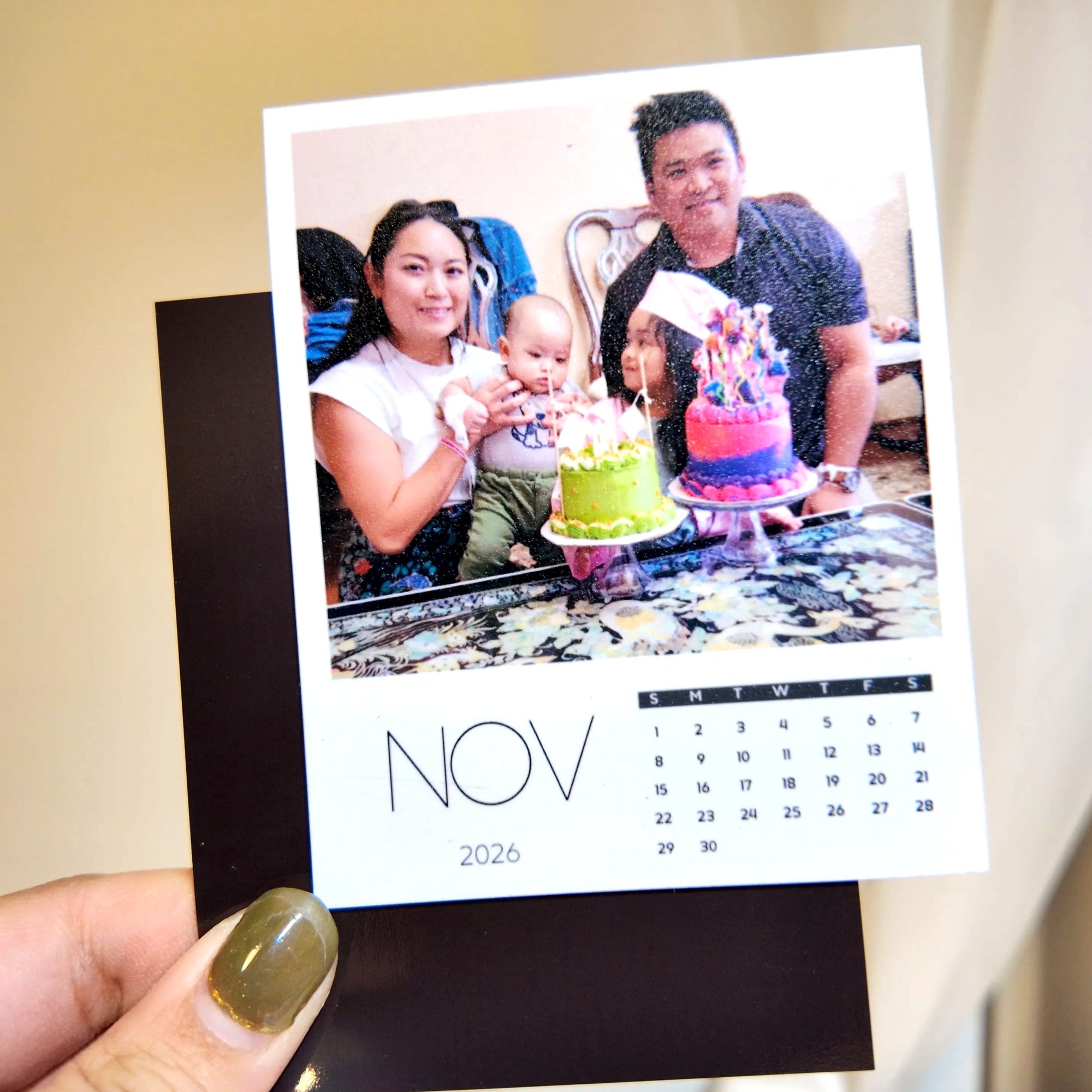 A person holding a photo of a family celebrating a birthday, with two cakes and a baby present.