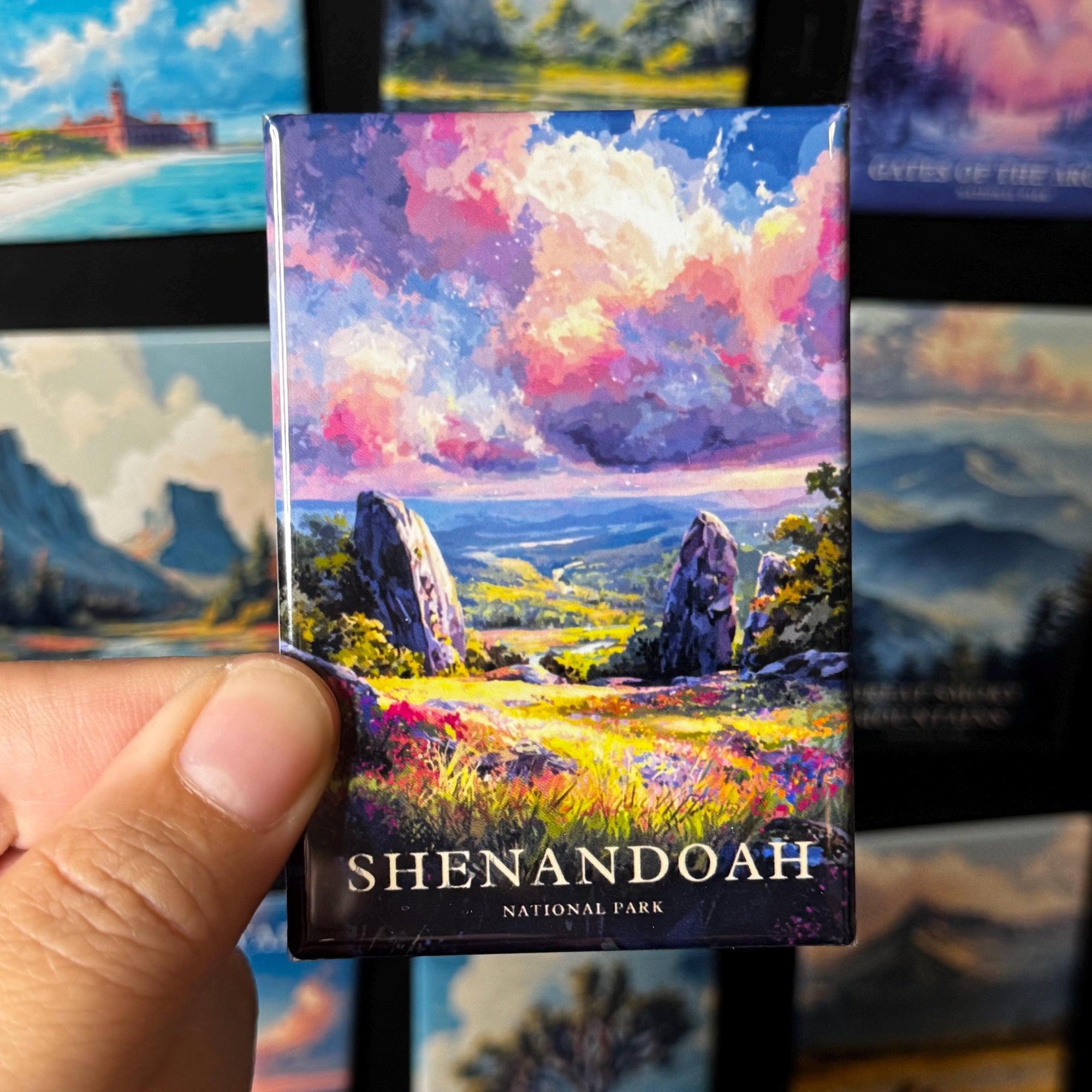 A hand holding a magnet with a colorful painting of a scenic landscape, featuring mountains, clouds, and a river.