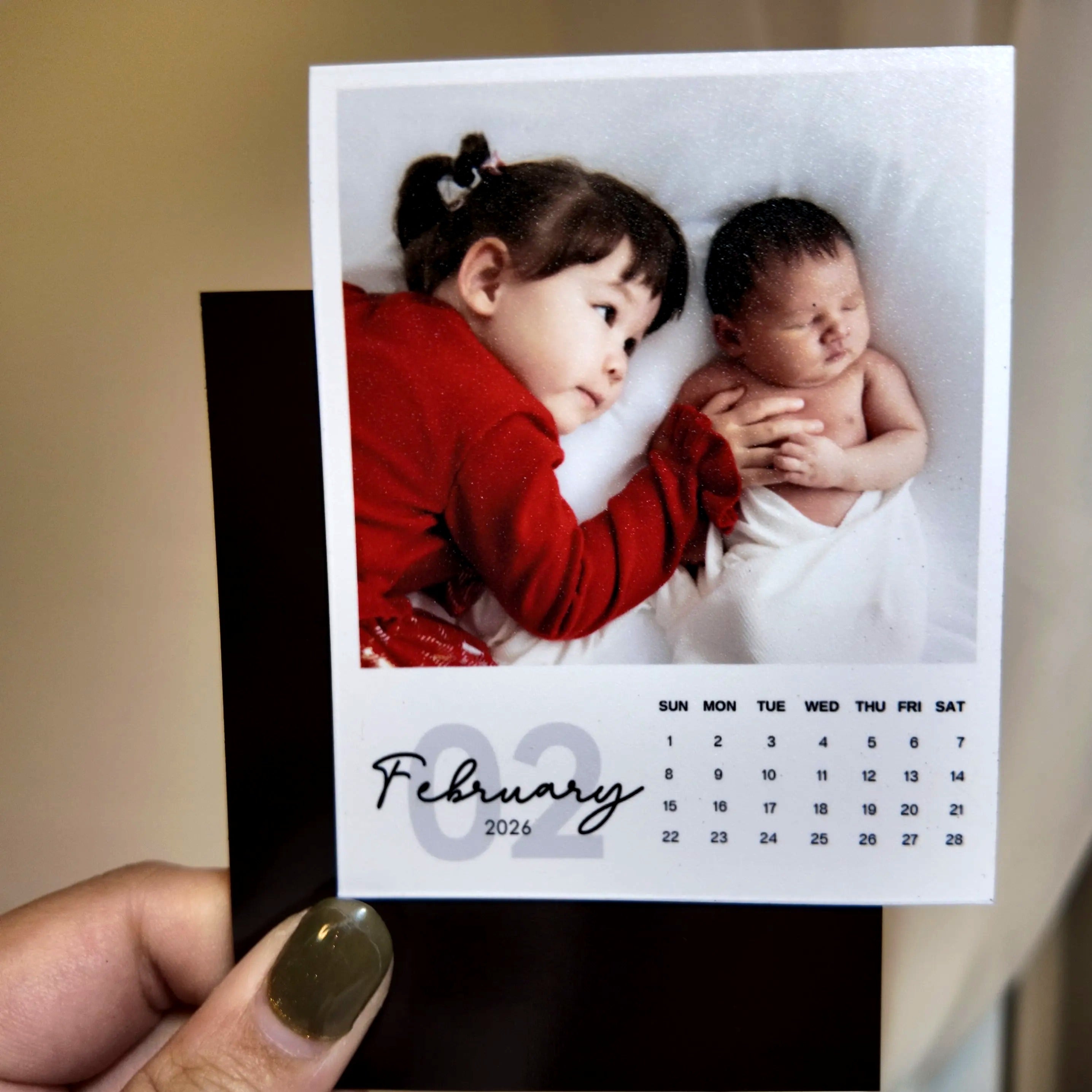 A person is holding a small photo album with a photo of two young children, one of whom is a baby, and the date "FEBRUARY 2026" printed on it.