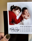 A person is holding a small photo album with a photo of two young children, one of whom is a baby, and the date "FEBRUARY 2026" printed on it.