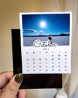 A person's hand holding a calendar for the month of October, with a scenic landscape visible in the background.